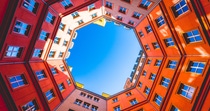 View of vibrant orange building walls from below, forming an octagonal courtyard. Blue sky visible above, creating a dynamic and geometric perspective. View of vibrant orange building walls from below, forming an octagonal courtyard. Blue sky visible above, creating a dynamic and geometric perspective.